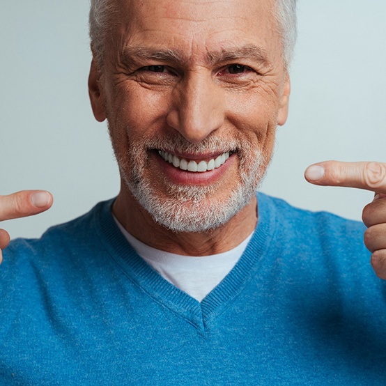 Man in blue sweater with white facial hair pointing to his denture smile